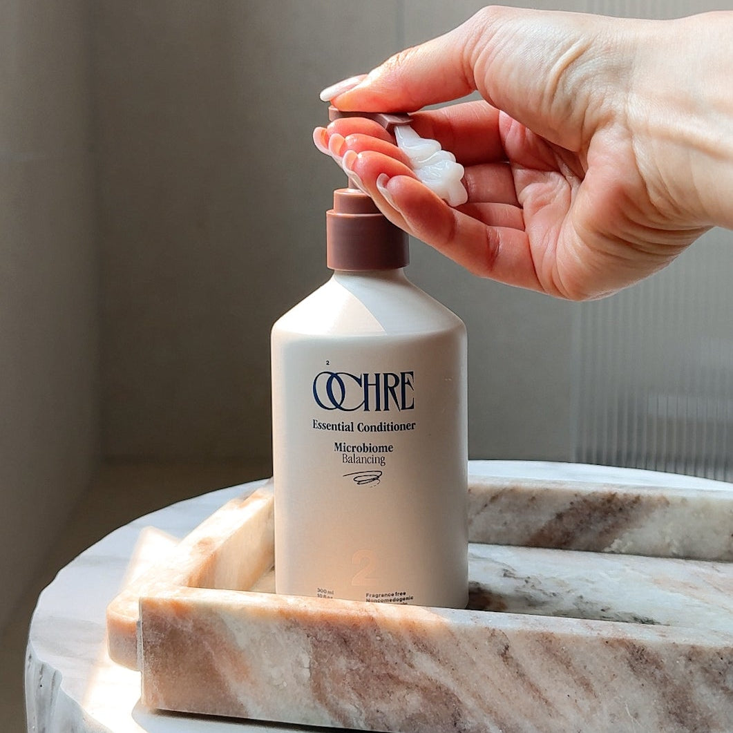 A hand dispenses white lotion onto a marble tray holding a bottle of O'HRÉ Essential Conditioner, set on a marble pedestal in a softly lit bathroom setting.