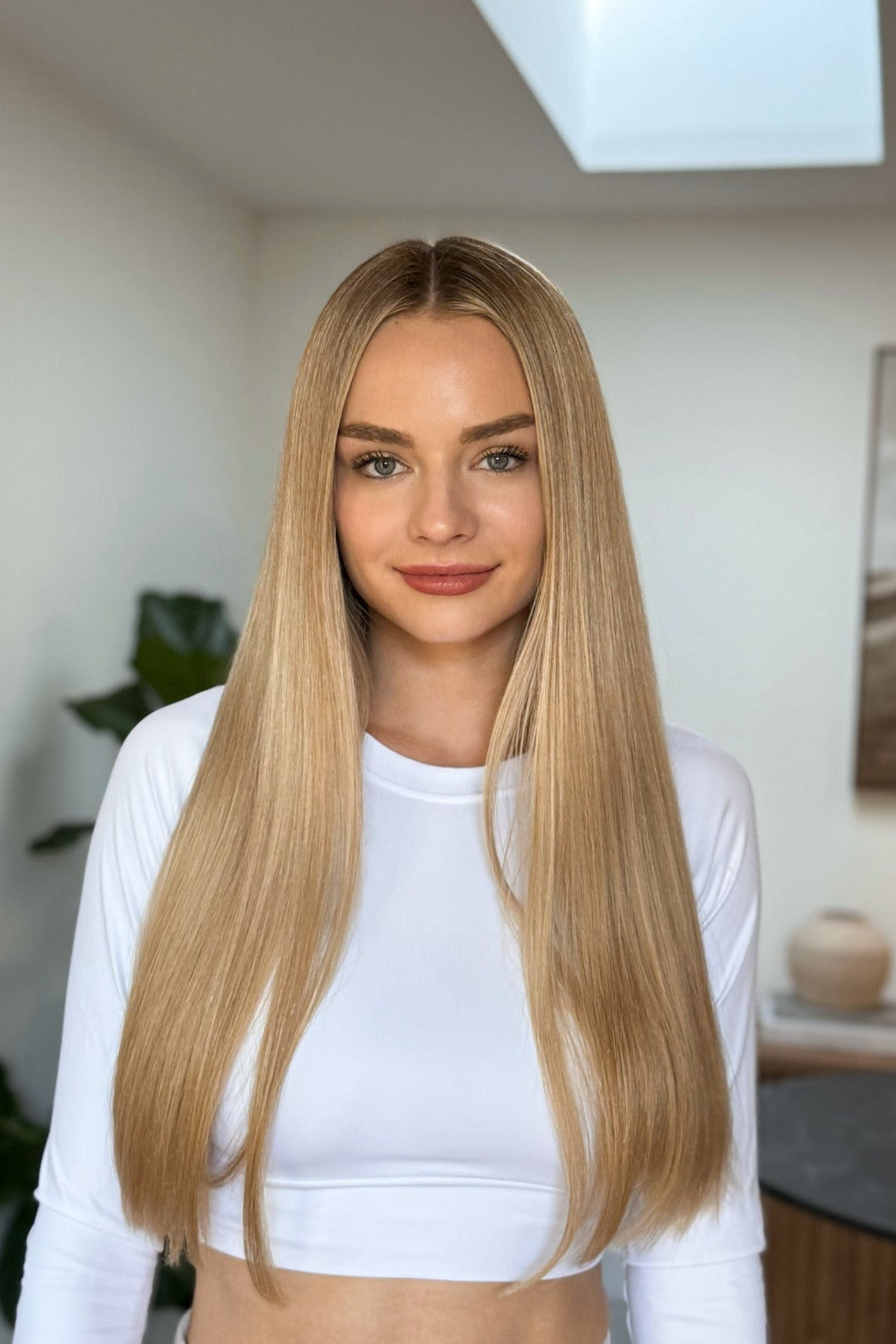 Woman with long blonde hair wearing a white top in a room with a plant and decor.
